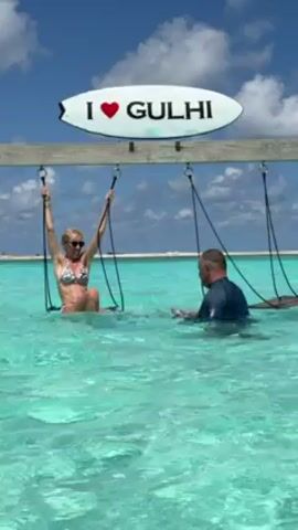 A woman in a patterned bikini and sunglasses sits on a swing in shallow turquoise water. A man in a dark wetsuit stands in front of her, holding what appears to be a camera housing. The swing hangs from a wooden frame with a surfboard-shaped sign that reads "I (heart symbol) GULHI". The sky is blue with puffy white clouds, and a white sandy beach is visible in the background. The woman is smiling and appears to be posing for the camera.