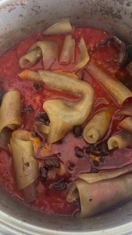 A close-up shot shows a silver pot filled with a reddish-orange sauce containing various cooked ingredients. The sauce appears to have a slightly oily sheen. There are several pieces of a pale, translucent ingredient that are curled or rolled, and some are cut into flat shapes. Dark brown, irregular pieces are scattered throughout the sauce as well. A gray slotted spoon is partially visible, being used to stir the contents of the pot. The pot itself is slightly scratched and shows signs of use.