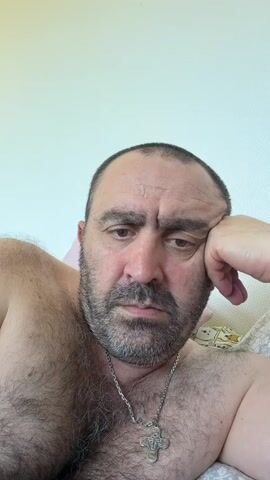 This is a close-up, first-person perspective selfie of a man lying down. He is shirtless, revealing dark hair on his chest and shoulders, and is wearing a silver chain with a cross pendant. The man has short, dark hair, a beard and mustache, and appears to be in his 40s or 50s. His right hand is resting on his cheek, and his expression is somber. The background is out of focus, but appears to be a bed with light-colored bedding and a white wall. The lighting suggests it is daytime. The overall mood is one of quiet contemplation or weariness. There is no visible text in the image.
