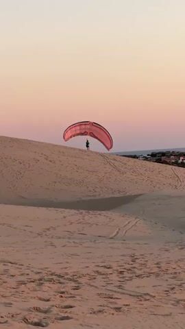 A paraglider prepares to launch from a sand dune at sunset. The paraglider's wing is a vibrant pink and red, taut in the air. The pilot, wearing a green shirt and dark pants, stands on the crest of the dune with arms outstretched, holding the control lines. The sand dune is covered in footprints and tire tracks, suggesting previous activity. The sky above is a soft gradient of pale pink and orange, indicative of the twilight hour. In the distance, a line of buildings peeks over the horizon. The overall scene conveys a sense of anticipation and adventure.