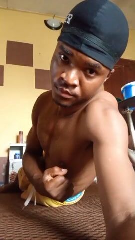The video shows a shirtless man wearing a black du-rag and yellow shorts. He is flexing his bicep and looking directly at the camera. He is lying on a patterned brown rug. The background features a wall with alternating brown and yellow squares, a shelf with items on it, and a small table with a blue bucket.