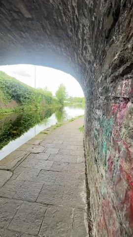 The video shows a view of a canal from inside a stone tunnel. The path is paved with gray rectangular stones. The left side of the canal has a grassy bank and some foliage. The right side of the tunnel wall has graffiti. The water reflects the vegetation on the opposite bank. In the distance beyond the tunnel entrance, more of the canal and some trees are visible.  The sky is overcast.