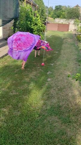 In an outdoor grassy area, a child wearing a pink shirt and shorts is practicing gymnastics. The child is holding a long, flowing purple scarf with pink floral designs. The scarf is attached to a wire that is strung between two poles. The child is moving the scarf in a circular motion around themselves. In the background, there is a fence, trees, and a building. The weather appears to be sunny.