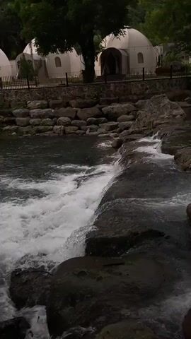 The video shows a small, rocky waterfall cascading into a pool of water. A row of dome-shaped tents is visible in the background, lined by a low stone wall and black fence. There is lush greenery and trees. Someone speaks in the background. The lighting is subdued.
