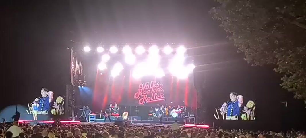 The video shows a live outdoor concert performance at night. The stage is brightly lit with numerous spotlights and has a large screen in the background displaying the text "Volks Rock'n'Roller" in a stylized font. On stage, a band is performing, with several musicians visible playing instruments like guitars and drums. A lead singer, dressed in a blue and gold outfit, is on the right side of the stage interacting with the audience. Two large video screens on either side of the main stage show close-ups of the lead singer. A large crowd of people is gathered in front of the stage, watching the performance. The overall atmosphere appears energetic and festive.