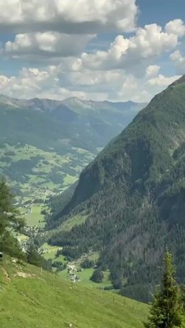 A drone shot captures a scenic mountain landscape with lush green valleys, forested slopes, and a winding road. The sky is partly cloudy with white cumulus clouds against a blue background. The camera moves from left to right, revealing more of the valley and distant mountain ranges.