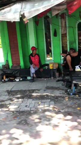 Two men are sitting in front of a green building. The man on the left is wearing a red shirt with black sleeves and a red hat. He is sitting on a bench and appears to be looking at the man on the right. The man on the right is wearing a dark shirt and is sitting on a bench as well. He is pointing towards the left. The building behind them is painted green with red stripes. There are various objects on the ground, including shoes, tools, and containers. The ground is paved with interlocking blocks. The lighting suggests it is daytime.