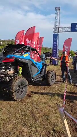 The media file shows a blue and black UTV (Utility Terrain Vehicle) with yellow suspension springs on a dirt track. The UTV is positioned on the left side of the frame, facing right. A person in an orange safety vest and a white cap is standing to the right of the UTV, holding a rope or cable attached to it. Another person is standing further to the right, looking towards the UTV.

In the background, there are several tall, red flags with white lettering, and a large metal structure that appears to be an archway or start/finish line for a race. There are also other people visible in the background, some near the flags and others further away. The setting appears to be an outdoor event, possibly a motorsports race or rally, given the UTV, the flags, and the start/finish line structure. The ground is covered in dirt and dry grass, suggesting a rural or off-road location. The sky is visible above with some clouds, and the lighting indicates it is daytime, likely late morning or afternoon.

Visible text on the flags includes "MAX" and "LYNX" and other smaller logos. The UTV itself has "RZR" written on its side. The start/finish line structure has several logos and text, including "START" clearly visible on the blue banner. The overall scene suggests an organized event taking place under a partly cloudy sky.
