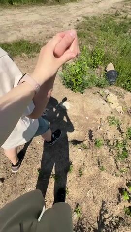 The video shows two people interacting outdoors. One person, seen from the waist down, is wearing light-colored pants and black sneakers. This person's shadow is cast on the ground. The other person, whose upper body is visible, is wearing a light-colored t-shirt, shorts, and black sneakers. This person has a watch with a pink band on their left wrist. Both individuals are holding hands and engaging in some form of hand gesture, possibly a handshake or a playful interaction. The environment appears to be a dirt area with sparse vegetation and shadows indicating a sunny day.