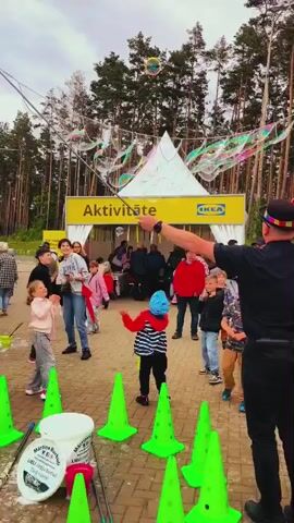 A man uses a long stick to create large bubbles, while children watch and play around him. He is wearing a black shirt, a black hat with colorful squares, and a black apron. A woman in a black t-shirt helps him. Many bubbles float in the air, reflecting various colors. A large white tent with yellow signs that read "Aktivitäte" and "IKEA" is visible in the background. Several people, including families and children, are gathered around. Tall trees line the background. The sky is overcast.