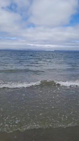 This is a video of a beach scene. The water is a deep blue, with gentle waves rolling onto a sandy shore. The sky is partly cloudy, with patches of blue peeking through. In the distance, the hazy outline of land can be seen. The overall mood is peaceful and serene. The location is Pier Road, Ireland. The time of day appears to be daytime, with bright but diffused light suggesting it may be overcast or the sun is low in the sky. There is no visible text in the video.