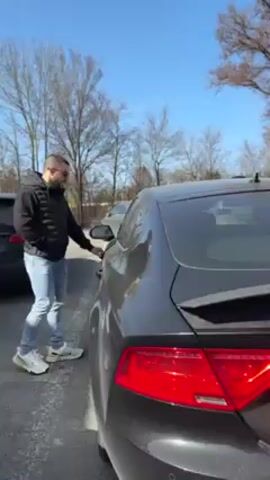 A person wearing a black jacket, blue jeans, and white sneakers is opening the driver-side door of a dark gray Audi A7. The car is parked in a parking lot on a sunny day. In the background, there are bare trees and a white box truck. The car has a German license plate that reads "GAP-YD 643". The rear spoiler is up. The person is facing away from the camera.
