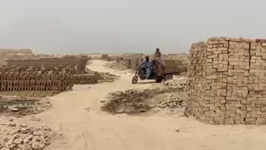 The scene depicts a dusty landscape with stacks of bricks in the foreground, middle ground, and background. Two men are riding a motorcycle-drawn cart filled with bricks on a dirt road. The sky is overcast and hazy, suggesting a dry, arid environment. The focus is on the transportation of bricks via motorcycle.