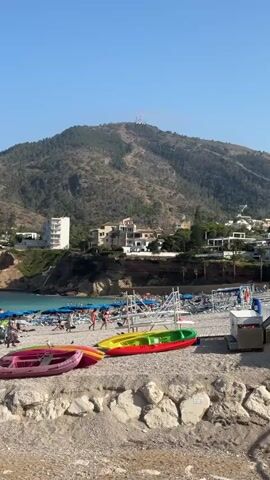 The video shows a crowded beach with people relaxing on lounge chairs under umbrellas, swimming in the ocean, and walking along the shore. In the foreground, several colorful kayaks and pedal boats are resting on the pebble beach. Behind the beach, there are buildings and a large, tree-covered mountain under a clear blue sky. The overall scene depicts a lively and sunny day at a popular tourist destination.