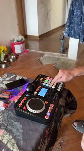 A person's hands are seen operating a DJ controller. The controller is black and has various buttons, knobs, and a jog wheel. The person is pressing buttons on the controller, and a small blue screen is visible. The controller is placed on a table covered with a floral-patterned cloth. In the background, there is a room with wooden floors and some unfinished construction work visible. A phone and some pet bowls are also on the floor near the table.