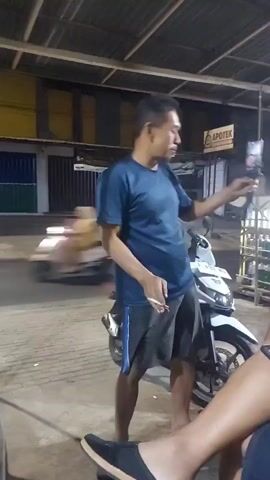 A man in a blue t-shirt and shorts holds a cigarette in his right hand while standing next to a white motorcycle. He is looking at a smartphone being held by another person, partially visible in the foreground, who is seated and wearing a blue jacket. A second man, wearing a black cap, is also seated and appears to be talking to the man in the blue t-shirt. They are under a roofed structure, possibly an outdoor seating area, with shops and a street visible in the background. The lighting suggests it is nighttime.