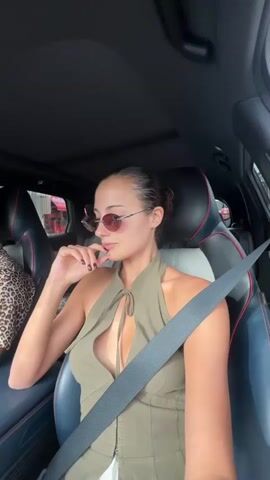 The video shows a woman sitting in the driver's seat of a car, wearing sunglasses, an olive green halter top, and a seatbelt. She is looking around and appears relaxed, with her hair pulled back. The car has black and grey leather seats with red accents. The passenger's seat next to her is partially visible, occupied by a person wearing a leopard print top. The car seems to be moving through a city, as glimpses of buildings and traffic can be seen through the windows. Music is playing in the background.