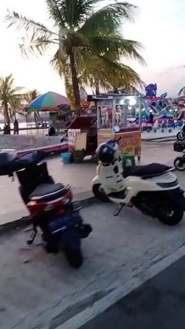 The video shows a street scene at dusk with several amusement rides and food stalls set up. In the foreground, there are parked motorcycles and scooters. Further back, there are colorful rides, including what appears to be a Transformers-themed attraction. Palm trees line the background, and the sky is a pale orange, suggesting sunset or sunrise. People are visible near the stalls and rides, but their actions are not clearly discernible due to the distance and low light. The overall impression is of a temporary amusement area preparing for or winding down operations for the day.