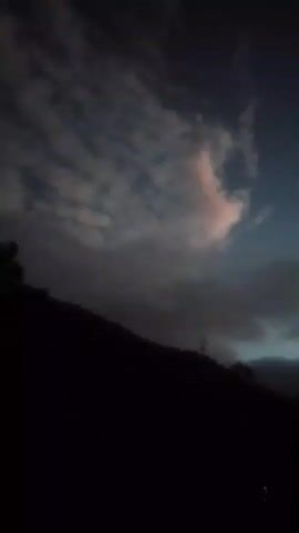 The video shows a dark, silhouetted landscape with a sky filled with clouds during twilight. The sky transitions from a dark blue at the top to a lighter shade near the horizon. Wispy clouds, tinged with pink and orange hues, are scattered across the sky. A bright, circular object, likely the moon or a star, is visible in the upper portion of the frame. The lower part of the image is dominated by the dark silhouette of a hill or mountain, with some indistinct shapes of trees or structures on its slope. There are no people or clear objects visible in the foreground. The overall mood is serene and captures the transition from day to night.