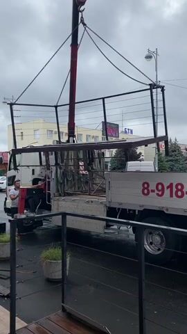 A crane is lifting a metal structure from the back of a truck. The structure appears to be a section of fencing or railing. A man is standing next to the truck, appearing to guide the process. The truck has the phone number 8-918-39-96 on the side. The scene appears to be outdoors on a cloudy day, with buildings and trees in the background.