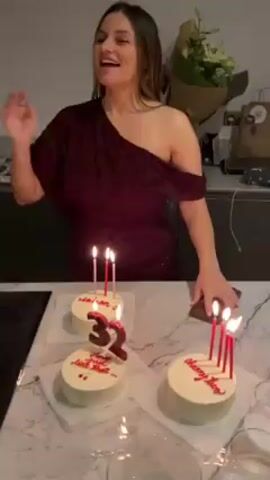 A woman celebrates her birthday in a kitchen. Three small, round cakes are on a countertop, each with white frosting and red lettering. Candles are lit in each cake. The middle cake has a "32" made of red fondant. The woman wears a maroon dress and takes photos of the cakes with her phone. A bouquet of flowers rests on the counter behind the cakes. The kitchen has modern cabinetry and lighting.
