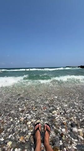 The video shows a first-person perspective of someone standing on a pebble beach with their feet in black flip-flops. The ocean is in front of them with waves rolling in. People can be seen swimming or playing in the water in the distance. The sky is clear and blue.