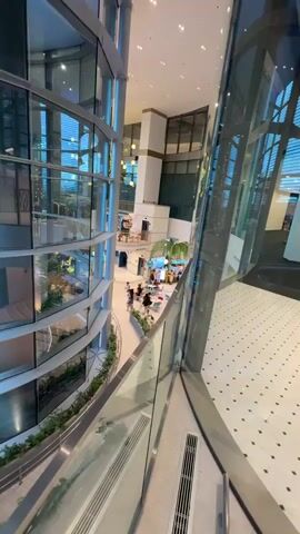 The video shows an interior view of a modern building with a curved glass facade. Several floors are visible, with a central atrium or open space. The lower levels appear to be a retail or entertainment area with small shops or kiosks and people walking around. Plants and greenery are integrated into the design, particularly on the lower levels and along the walkways. There are tiered seating areas or staircases. The lighting suggests it is either dusk or evening, with artificial lights illuminating the interior and creating some atmospheric effects, including purple spotlights on the lower level. The perspective is from a higher vantage point, looking down and across the building's interior.