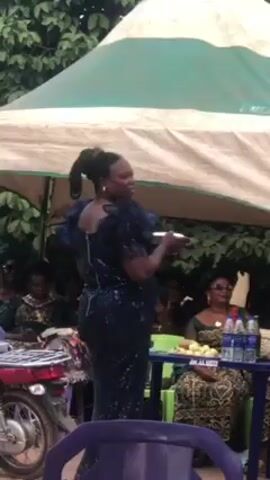 A woman in a dark blue sequined dress with ruffled sleeves stands speaking to an audience gathered under a white and green tent. She holds a small, flat object in her hand, possibly a tablet or book. The audience sits at tables, some are taking photos. A motorcycle is parked to the left of the woman, and scattered currency is visible on the ground. A sign on the table in front of the audience reads "AKALUGU CLUB". The setting appears to be an outdoor event.