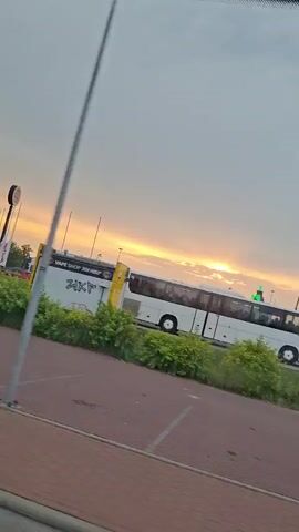 The video shows a moving perspective from a vehicle, looking out at a landscape during sunset. In the foreground, a blue hatchback car is partially visible, its rear end facing the camera. Beyond the car, a line of green bushes and trees is present, with a red-and-white sign partially obscured by vegetation. In the middle ground, a white tour bus with its door open is parked. Further in the background, a bright orange sun is visible through clouds, casting a warm glow across the sky. Several tall light poles are scattered throughout the scene, and structures that appear to be buildings and a Ferris wheel are visible in the distance. The overall impression is of an urban or suburban environment at dusk.