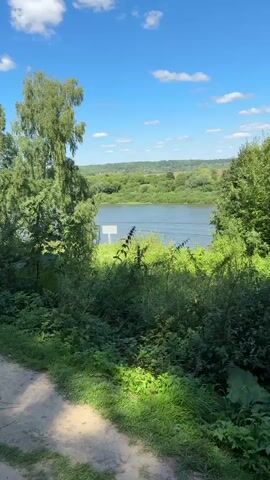 A sunny day with a blue sky and white clouds. A wide river is visible in the background, with green trees and hills on the opposite bank. In the foreground, there is a dirt path and lush green vegetation. A white sign is partially visible in the river.