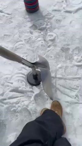 The video depicts a snow-covered area, possibly a ski slope. In the initial frame, there's a snow shovel with a black base, along with stacks of colorful rings forming cone-like shapes, scattered on the ground. Footprints are visible in the snow. The camera then pans to show a ski slope marked by blue and white flags. In the distance, a mountain with trees is seen, along with a ski lift. The sky is overcast with a few spots of sunlight. The video ends by showing an expansive snowy field with a skier.