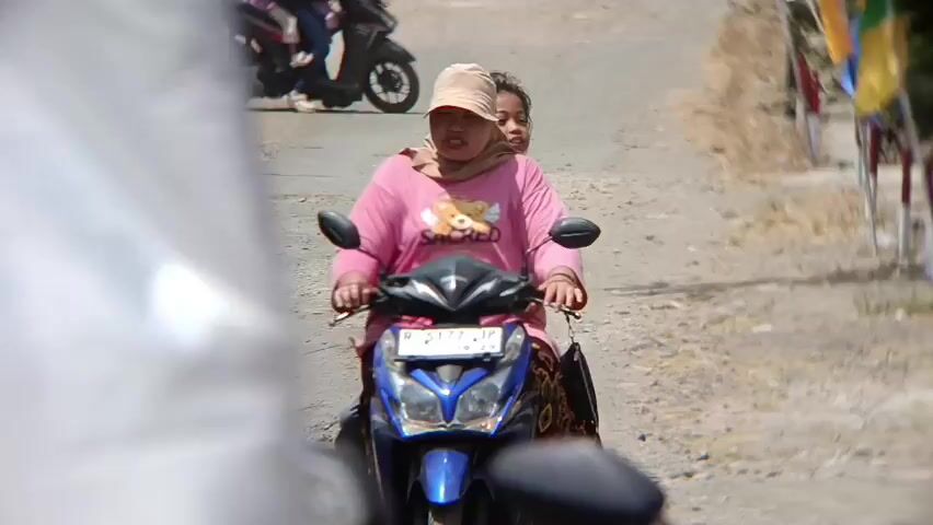 A woman wearing a pink long-sleeved shirt, a light-colored headscarf, and a hat rides a scooter on a dirt road. She has a child behind her. In the background, another person rides a scooter. There are flags on poles on the right side of the road. The road is dusty and appears to be in a rural area.