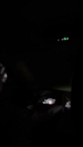 The video appears to be shot from inside a car driving at night. The interior is dark, with the dashboard and steering wheel faintly visible. The view through the windshield is limited due to the darkness, though some blurry shapes are discernible. The general environment appears to be outdoors.  There is audio of speech, but its content is not described.