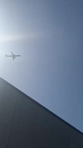 The video shows a plane in flight against a clear blue sky, partially obstructed by a gray wall in the bottom left corner. The sun shines brightly in the upper left, creating a glare. A contrail from another plane streaks across the upper right of the sky. The aircraft in focus appears to be a passenger plane, with a visible fuselage and wings. It is angled slightly, suggesting it might be turning or approaching. The environment appears to be outdoors, possibly near an airport or other aviation-related structure.