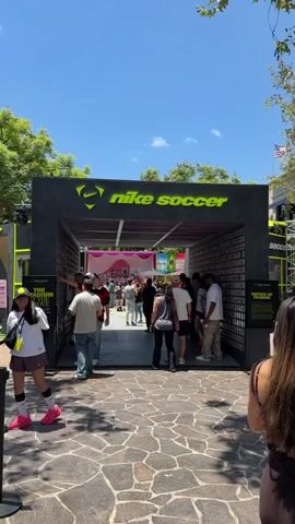 A group of people are gathered in front of a black archway that reads "NIKE SOCCER" in neon green. The archway has a grid-like pattern on its sides. Beyond the archway, there is a pink backdrop with decorations, and flags are visible. Some people are walking through the archway, while others are looking at displays on the walls. The ground is paved with stone. The scene appears to be outdoors, with trees and a blue sky visible.