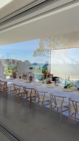 From behind a clear wall that reflects a bright outdoor scene, we see a formally-set dining table and two men preparing for an event. The long table is covered with a white cloth, place settings, and floral arrangements. The chairs are a light wood with woven seats. One man, wearing a dark shirt, is standing near the middle of the table, seemingly arranging items. The second man, wearing a white shirt and a mask, walks slowly in the background. Through the windows we see a sunny day, ocean views, greenery, and a dotted art piece. The space feels elegant and well-prepared.