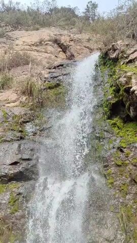This is a vertical video of a waterfall cascading down a rocky cliff face. The rocks are varied in color and texture, with patches of moss and vegetation growing on them. The water is white and frothy as it falls, creating a mist that partially obscures the rocks. The bottom of the waterfall shows water pooling around rocks before flowing away. The overall scene is natural and rugged, suggesting an outdoor, wilderness setting.