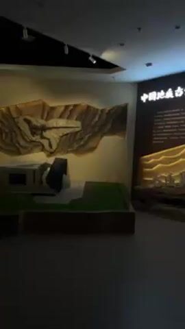 The video depicts the interior of a museum or exhibition hall. The scene is dimly lit, but spotlights illuminate the exhibits. There's a large wall display on the left with a relief sculpture depicting geological formations, possibly related to fossil finds. A model of what seems to be a structure sits in front of this display. On the right, another wall display features illuminated text and graphics, alongside portraits of individuals. The text appears to be in Chinese. The floor is smooth and grey. The overall ambiance suggests a scientific or educational setting.