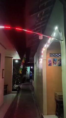 The video shows a narrow alleyway at night, illuminated by strings of red and white LED lights. The lights are strung across the top of the alley and are also arranged in a star shape on the wall of one of the buildings. The walls of the buildings are painted in light colors, and there are some decorative flags visible in the distance. The ground appears to be paved with concrete. The overall impression is one of festive or celebratory lighting.