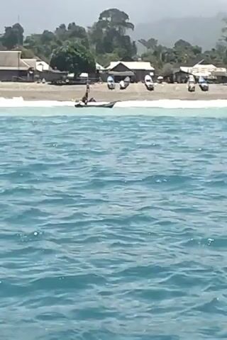 The video shows a body of water, likely the ocean, with a small village visible in the background. The village appears to be situated on a beach with several houses and boats. In the water, a small boat with at least two people on board is visible. The water is choppy with some waves, and the sky is overcast. The overall scene is one of a coastal community, possibly engaged in fishing or other marine activities.