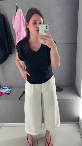 A woman stands in a dressing room, taking a mirror selfie with her phone. She is wearing a black v-neck t-shirt and loose, white, wide-leg pants. She has on black thong sandals. Her hair is pulled back into a messy bun. In the background, clothes hang on a rack, and a grey bench is visible. The walls are a textured grey. The woman shifts her pose and smiles at the camera.
