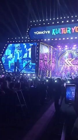 A concert audience is silhouetted in the foreground, holding up their cell phones to record the stage. The stage features a large screen displaying the band playing on stage. The stage itself is brightly lit with purple and white lights and features a circular design with the letters "K" and "&" in it. A large sign at the top reads "TÜRKİYE KULTUR YOLU FESTIVALI".