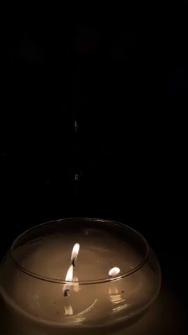 The video shows a close-up of a large, round, white candle with three lit wicks. The candle is in a clear glass bowl and is placed on a table. In the background, a vase of flowers, primarily pink and white tulips, is visible. The room is dimly lit, with the candlelight illuminating the foreground. The camera slowly zooms out, revealing more of the table and the flowers. The overall mood is serene and peaceful.