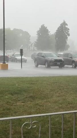 The video captures a heavy rainstorm with reduced visibility. Several cars are parked and driving on a wet road, with water splashing up from the tires. Trees and greenery are visible in the background, partially obscured by the rain and mist. The scene appears to be an outdoor parking lot or street.