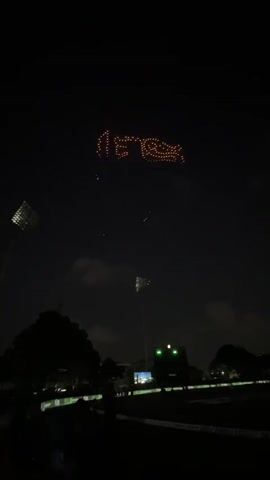 A drone light show displays the Sri Lanka Cricket logo in the night sky above a stadium. The logo, a lion rampant, is formed by orange lights. Below the lion, the words "Sri Lanka Cricket" are spelled out in white lights. The stadium is visible in the lower portion of the frame, with spotlights and a flag pole. Silhouettes of trees and people are visible in the foreground.