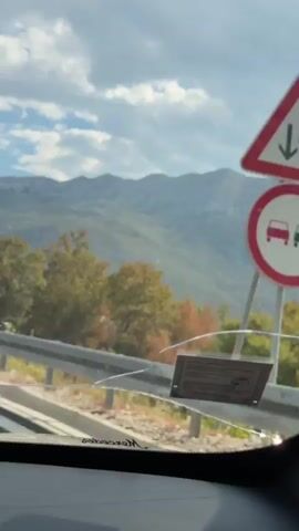 The video shows a view from inside a vehicle, looking out at a mountainous landscape. The sky is blue with scattered clouds. There is a guardrail in the foreground, and trees with autumn foliage can be seen. A wire fence with metal posts is visible behind the guardrail. The vehicle appears to be moving along a road.