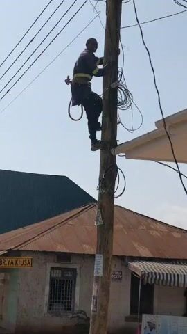 Here is a description of the video:

A person wearing dark clothing, a safety harness, and climbing spurs is ascending a utility pole. The pole is topped with various electrical and communication wires, and a box-like device. Below the pole are buildings with corrugated metal and white awnings. A sign on one of the buildings reads "BR.YA KIUSA." The sky is a pale blue. Sounds of passing traffic can be heard.