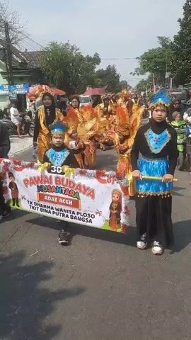 A parade is taking place on a sunny day on a street lined with trees and buildings. In the foreground, a banner reads "PAWAI BUDAYA NUSANTARA ADAT ACEH" in large, colorful letters. The banner is held by children in traditional costumes. To the right of the banner, a child wearing a blue and black costume with a headdress walks forward, holding a pole. To the left, other children in similar costumes are visible. Behind the children holding the banner, a group of people are walking in the parade, many dressed in elaborate gold and red costumes, some with feathered headdresses. Some individuals are carrying umbrellas or balloons. Vehicles, including motorcycles and cars, are visible in the background on the street. The overall atmosphere appears festive.