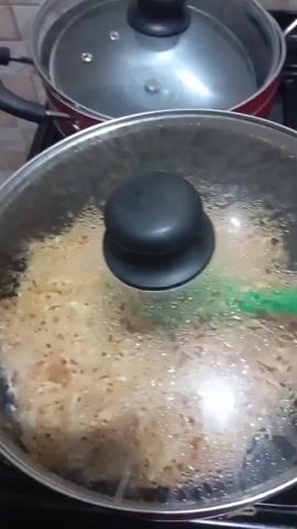 The video shows a close-up shot of a hand lifting a glass lid from a pan of spaghetti. The spaghetti appears to be cooked with some red sauce and possibly other ingredients, indicated by small red pieces mixed in. A green spaghetti server is visible within the pan, resting on top of the noodles. In the background, another pot with a lid is partially visible. The setting seems to be a kitchen, likely in Obada, Nigeria, given the provided context. The lighting suggests it might be daytime, though it's difficult to determine definitively. The overall impression is one of food preparation. There is no visible text in the video.