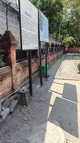 The video pans across an outdoor archaeological site with ancient brick ruins. In the foreground, a dirt path leads to a gravel-covered area with a few small plants. To the left, a brick wall is visible, with two information signs mounted on poles in front of it. A green trash can is next to the signs. The ruins, constructed of red brick, are situated further into the site, with a small, open-air structure housing a statue or monument. Trees and foliage surround the area, and the sky is clear and blue.